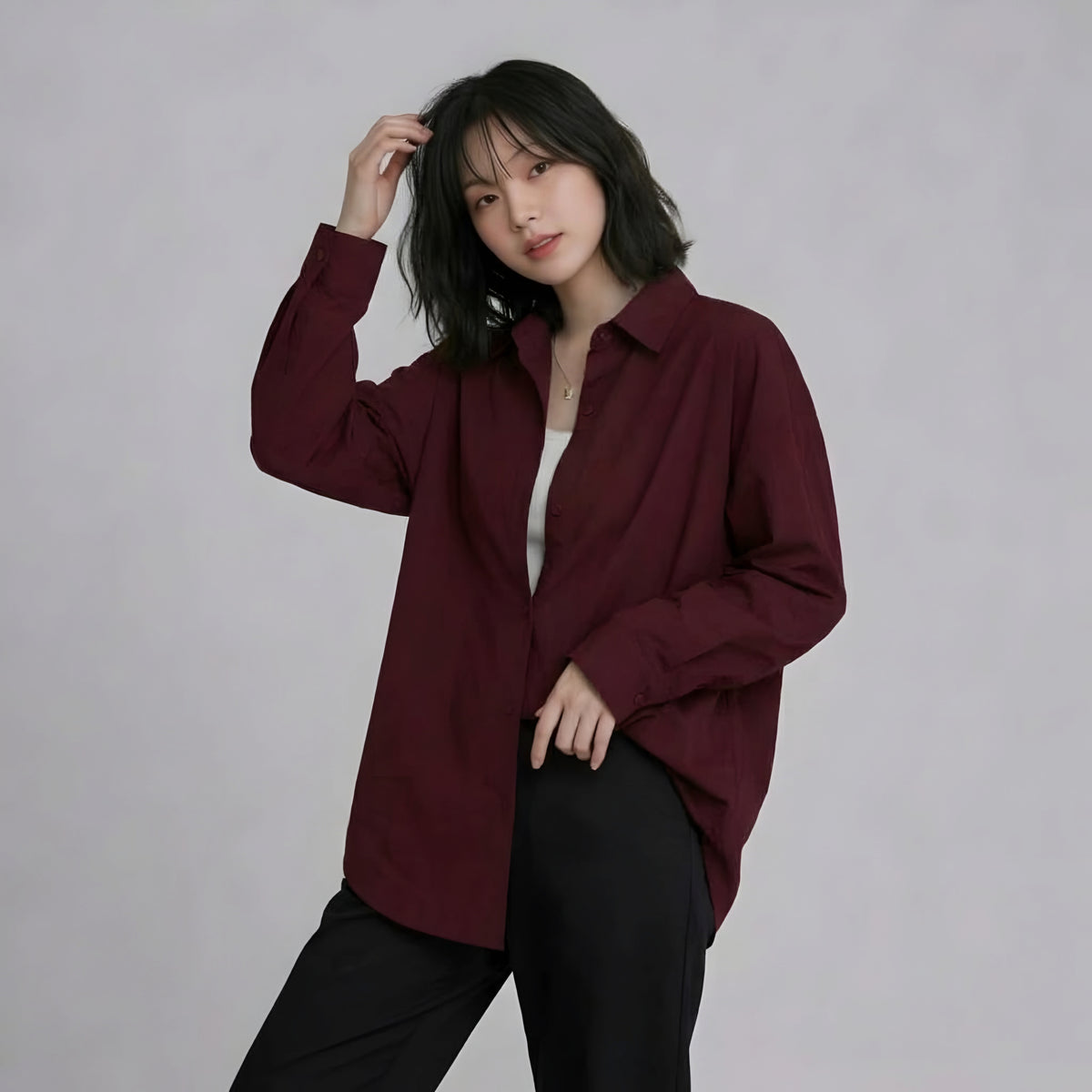 A woman modeling a long-sleeve button-up shirt in deep burgundy, worn open over a white top and black pants, against a plain light gray studio background.