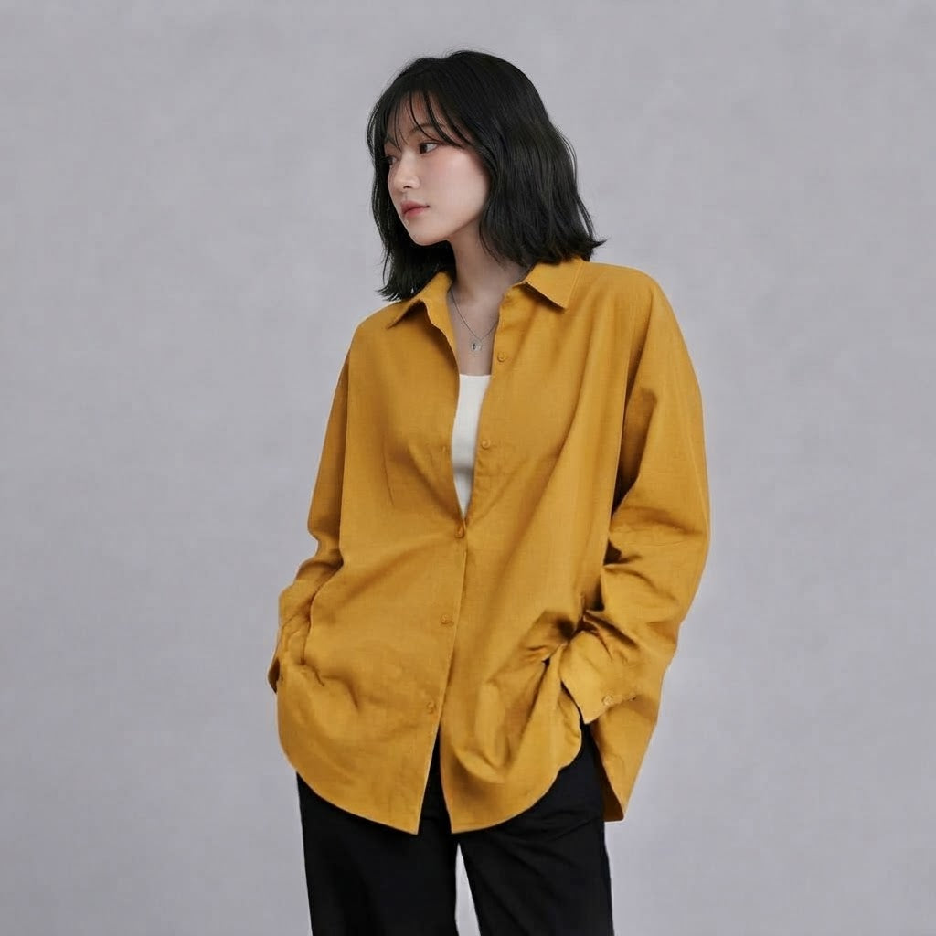 A woman wearing an oversized mustard yellow linen button-up shirt over a white top and black pants, standing against a textured gray studio background.