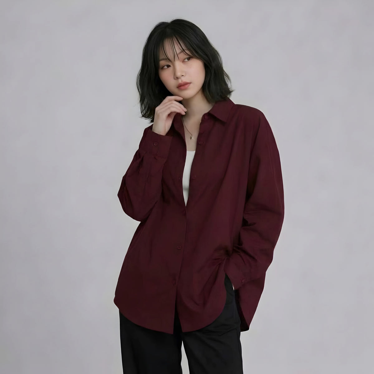 A woman with short dark hair wearing an oversized burgundy button-up shirt over a white top and black pants, standing against a light gray studio background.