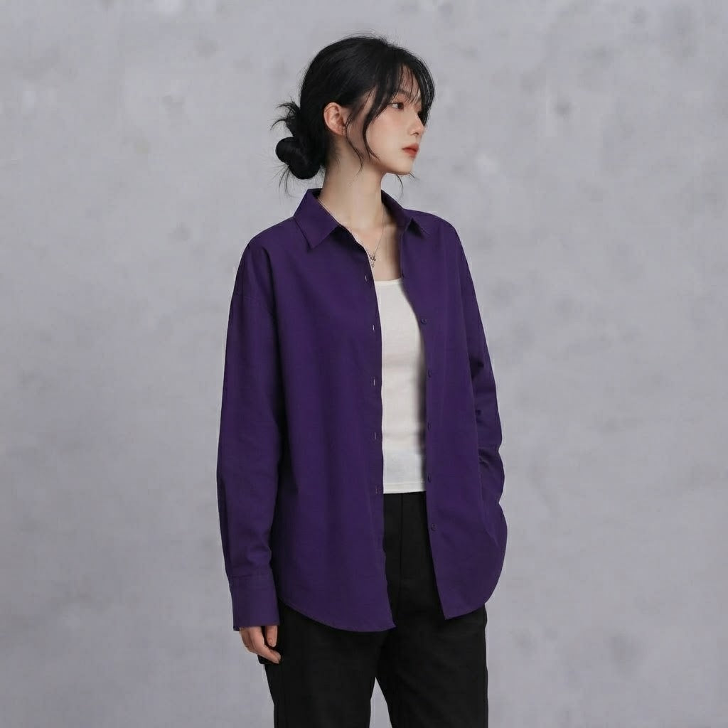 A woman wearing a long-sleeve purple button-up shirt over a white top and black pants, standing against a textured gray wall.