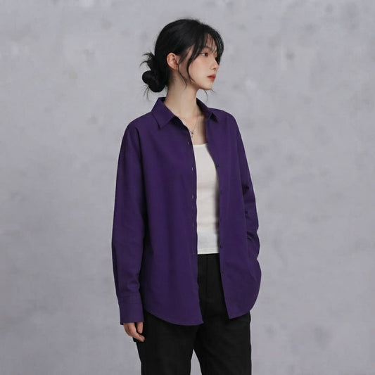 A woman wearing a long-sleeve purple button-up shirt over a white top and black pants, standing against a textured gray wall.