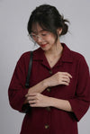 A woman wearing a textured burgundy button-up shirt with a collar and three-quarter sleeves, styled with a black shoulder bag and clear-framed glasses against a plain gray background.